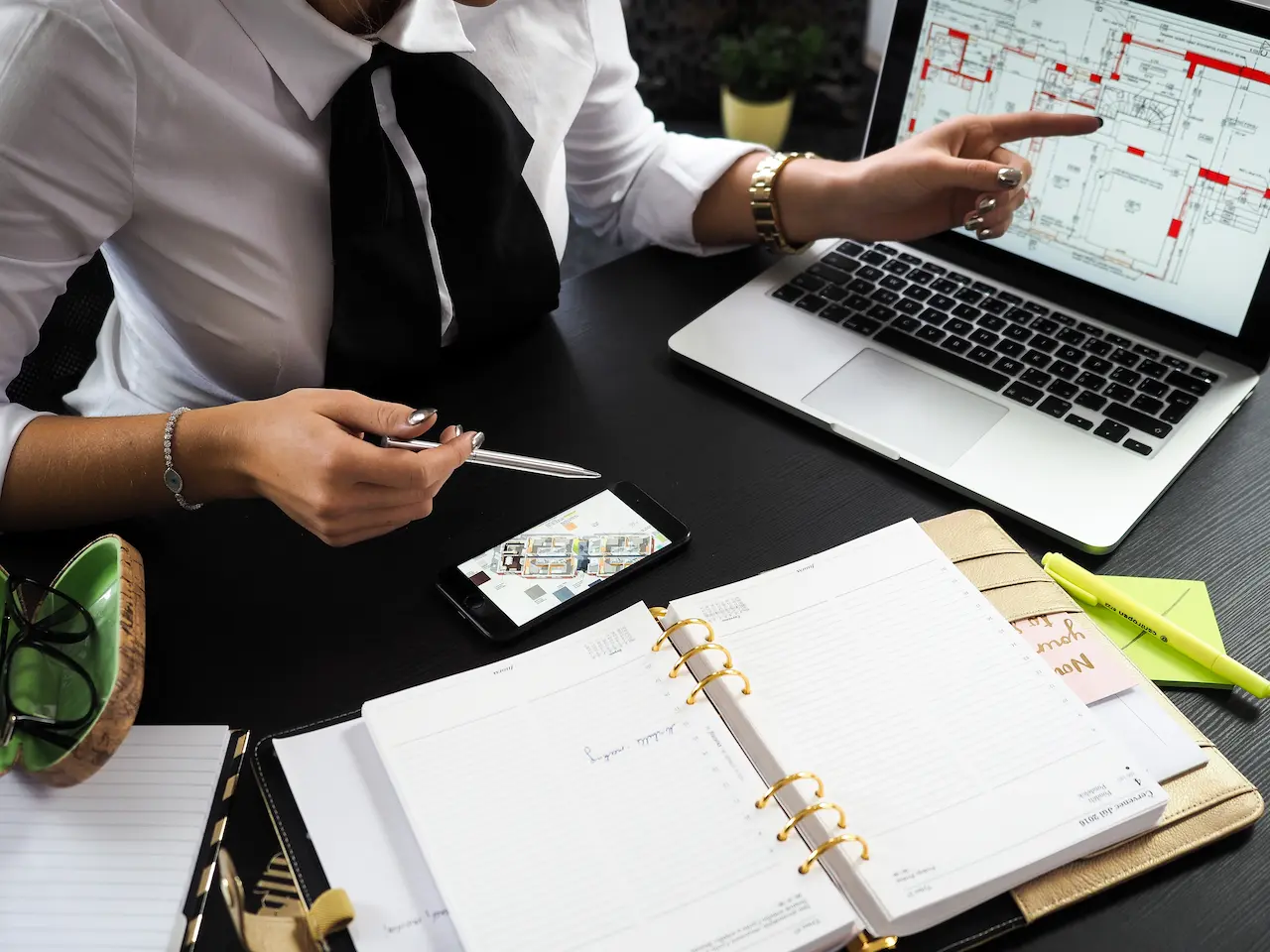 Person pointing at laptop screen with architectural plans, surrounded by an open planner, smartphone, and stationery on a black desk.