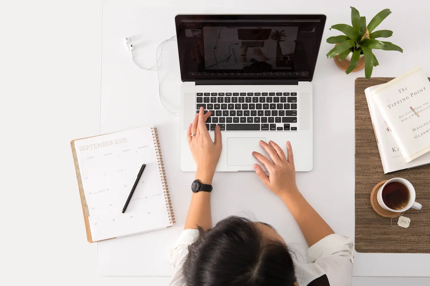 Person using a laptop at a desk with a calendar, plant, book, and cup of coffee.