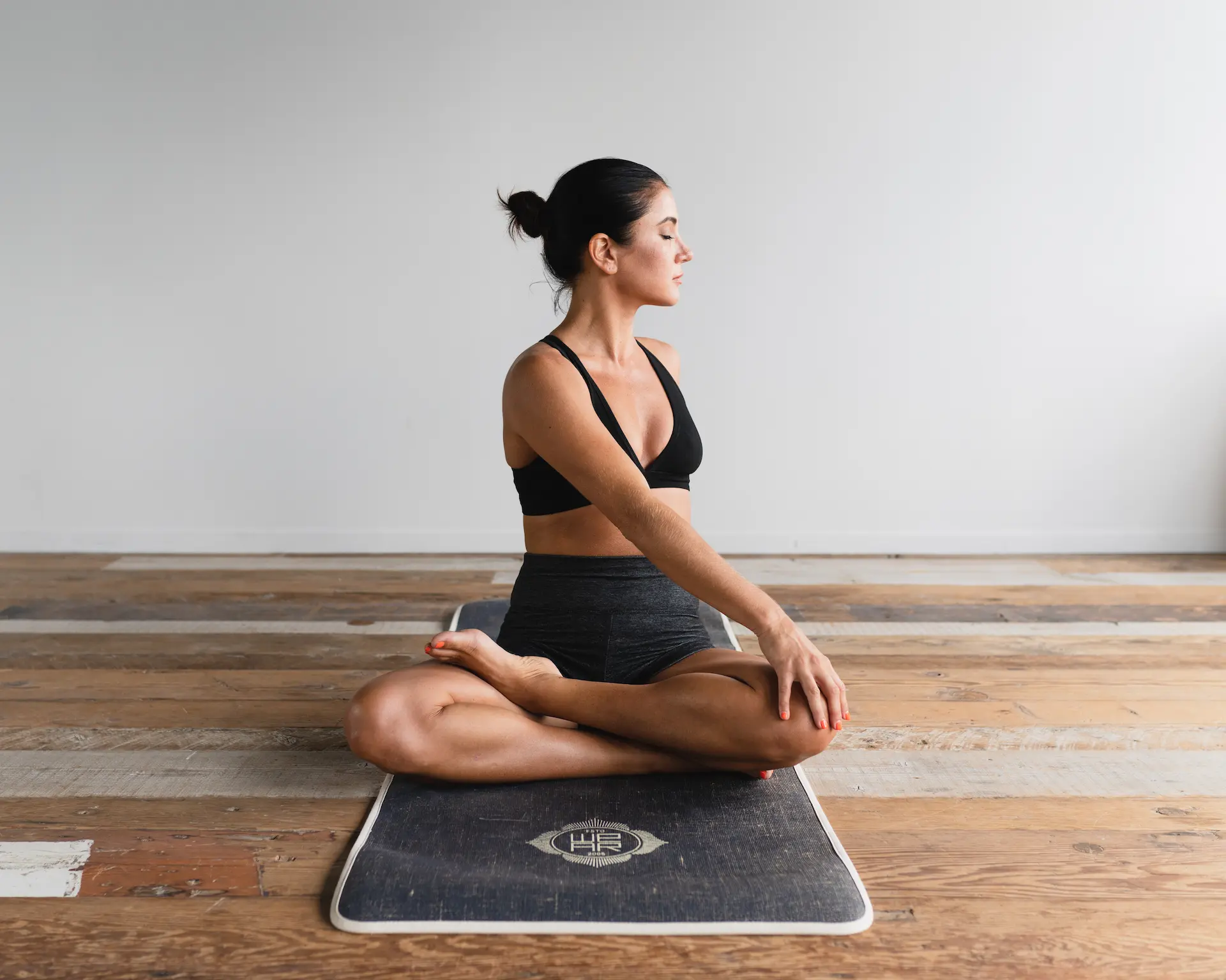 A woman in athletic wear sits cross-legged on a yoga mat, performing a seated twist pose in a bright, minimalist room.