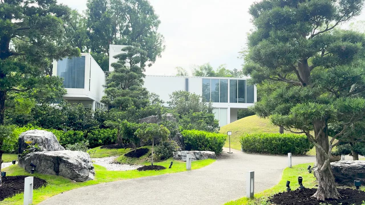Modern white building surrounded by lush greenery, trees, and a winding gravel path in a landscaped garden.
