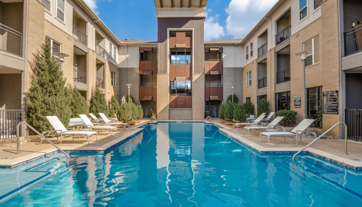 Outdoor swimming pool surrounded by lounge chairs and greenery, set in the courtyard of a modern apartment building under a blue sky.