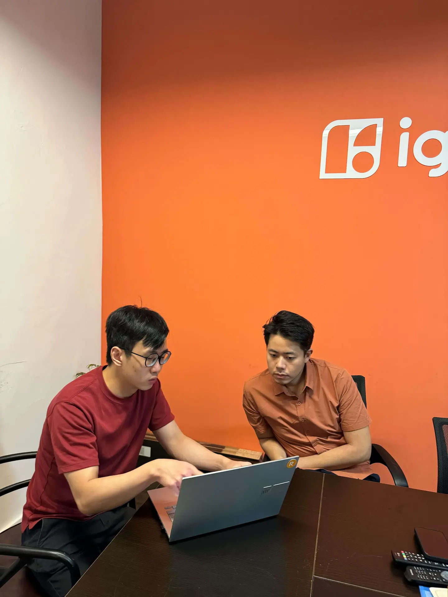 Two people are sitting at a table in an office, looking at a laptop. The wall behind them is orange with a logo.