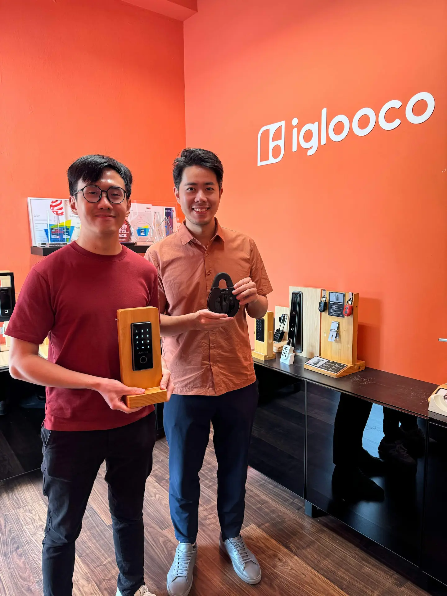 Two people standing in front of an orange wall with the "iglooco" logo, holding smart locks on wooden stands.