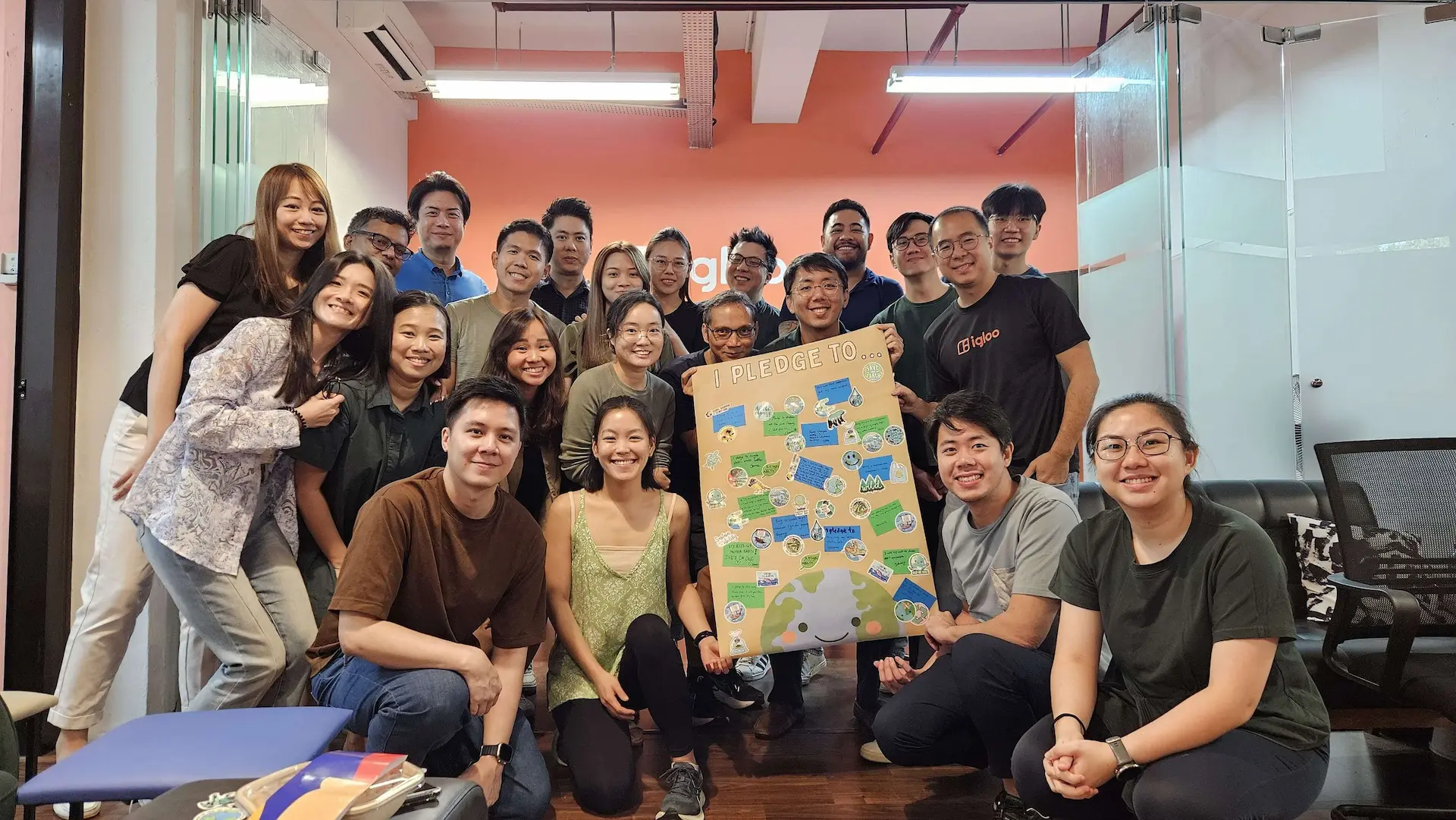 A group of people smiling and posing indoors, holding a colorful pledge board with various notes and stickers.