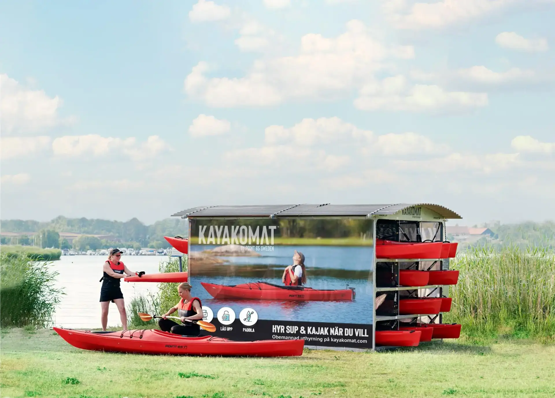 Self-serve kayaks lined up on a metal rack beside the water at a paddle sports hire site
