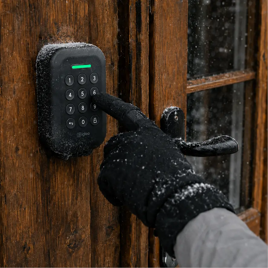 A gloved hand entering a PIN on a snow-covered igloo Lockbox fixed to a timber door