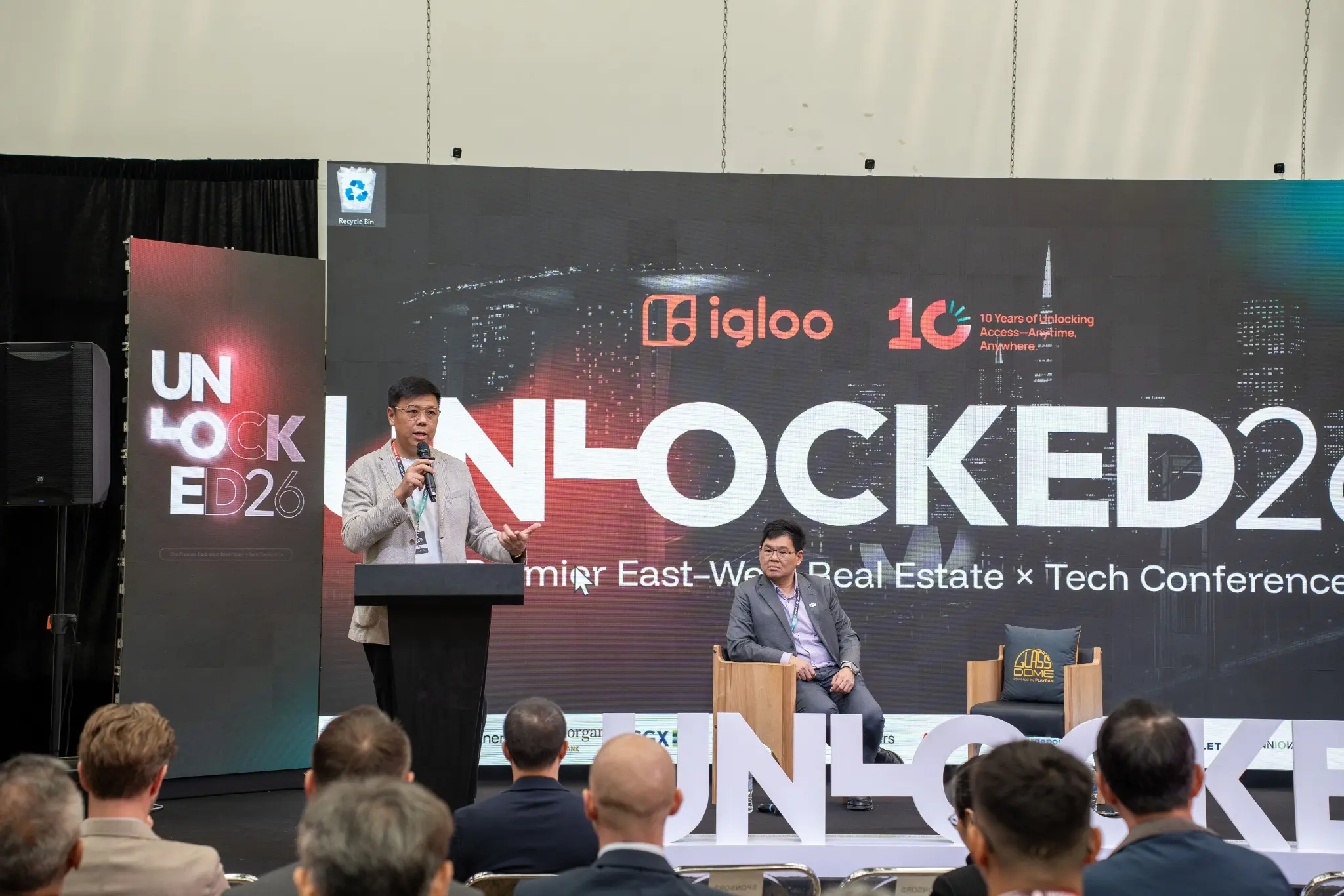 Speaker at the podium on the UNLOCKED26 main stage beside a seated guest