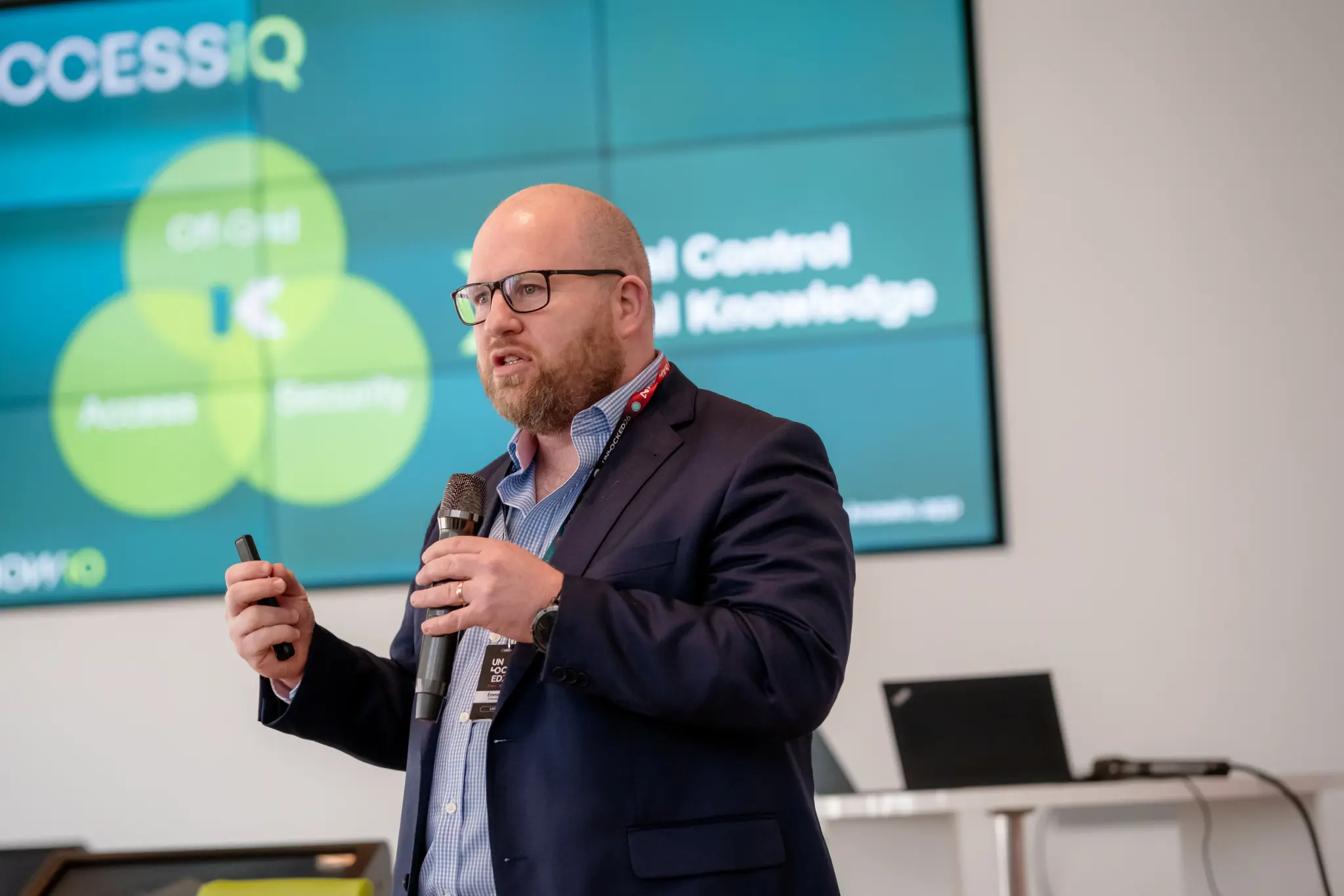 Speaker presenting Access IQ on stage with a microphone and clicker at UNLOCKED26