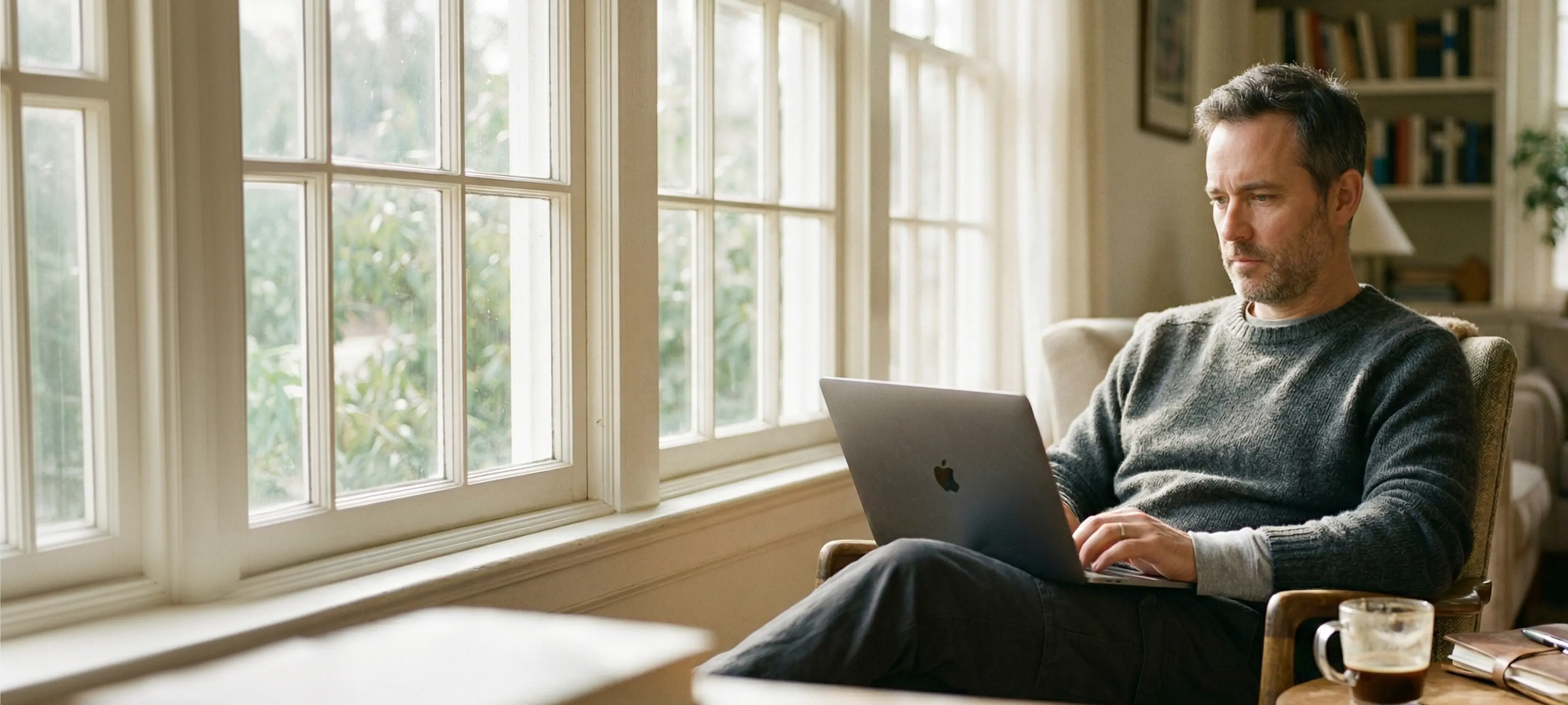 A property manager in a grey sweater sitting in an armchair in a sunlit living room, working on a laptop with a coffee beside him
