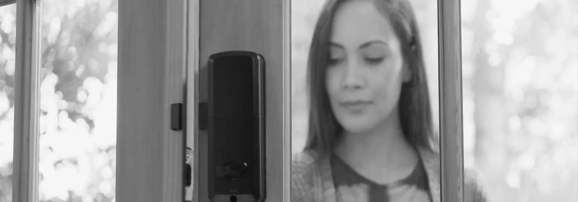 A woman behind a glass-panelled door fitted with the rear unit of an igloo smart deadbolt, in black and white