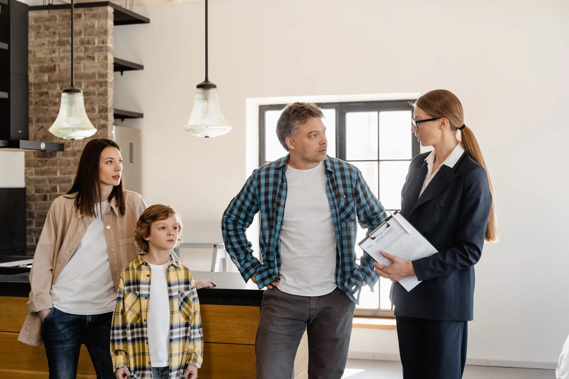 A family of three talks with a real estate agent in a modern kitchen. The agent holds a clipboard, and the family looks attentive.